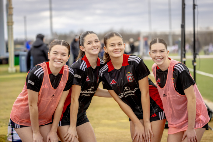 Four female soccer players pose on a field in black uniforms and pink vests on a cloudy day