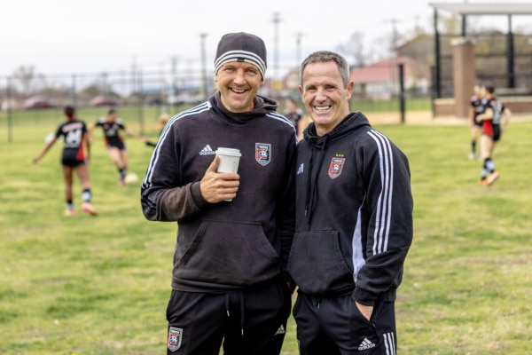 Two coaches smiling on a soccer field with players in the background