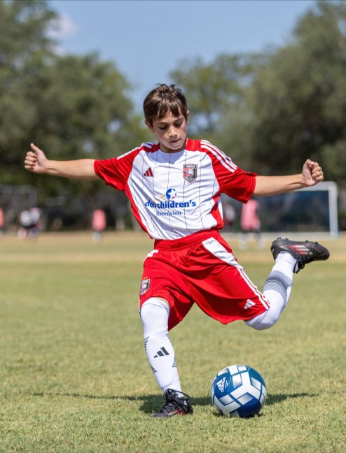 Young soccer player in red and white uniform kicking ball on grassy field.