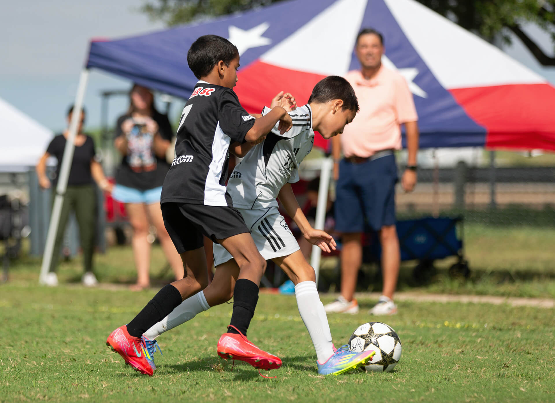 Two young boys playing soccer on a sunny day under a tent with spectators nearby