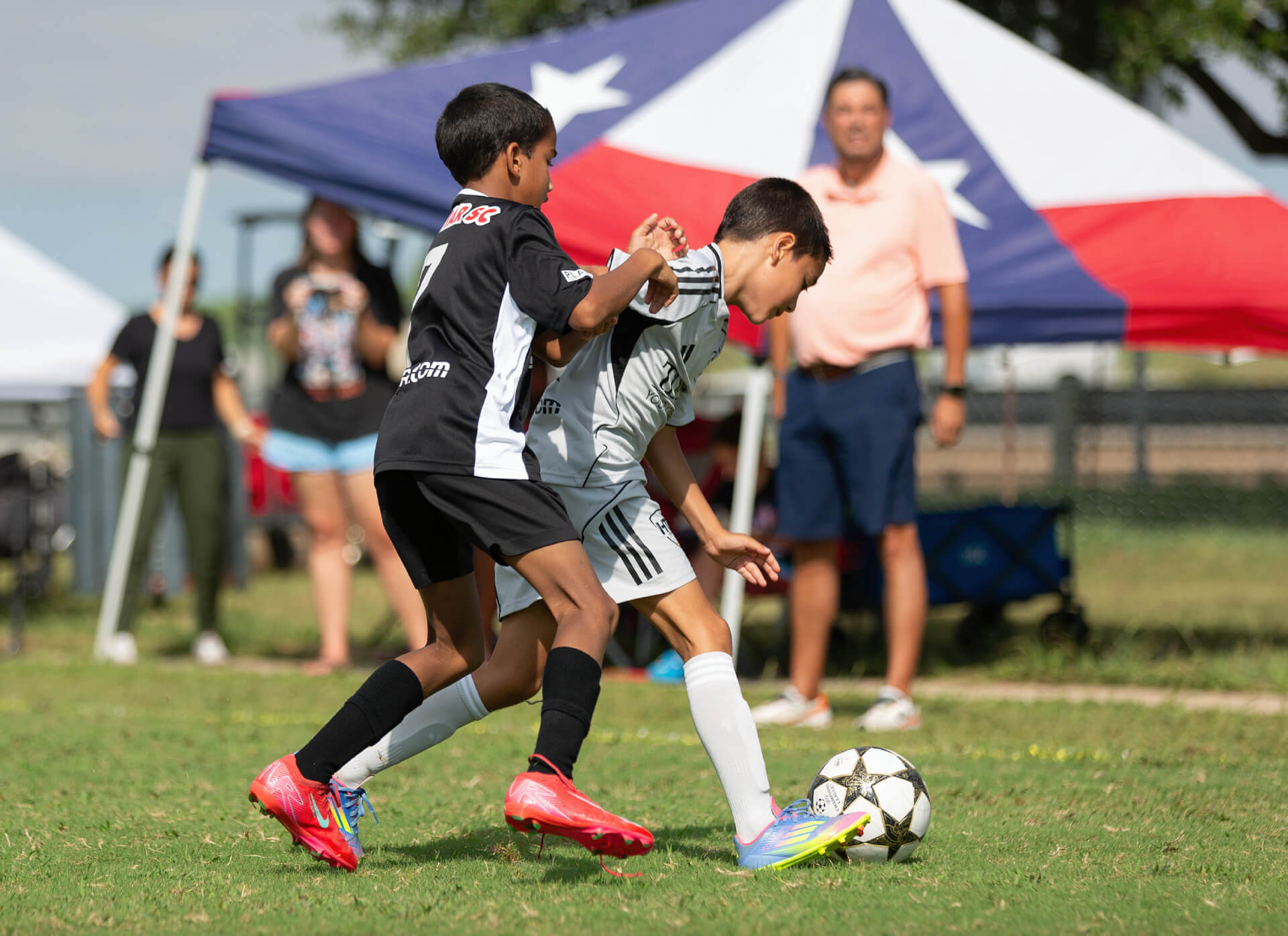 Kids playing soccer under a tent during a sunny day match.