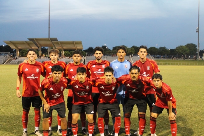 Soccer team in red uniforms posing on a grass field at dusk with a stadium structure in the background