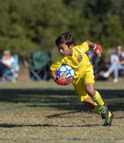 Young soccer goalie in yellow uniform diving to catch ball on grassy field
