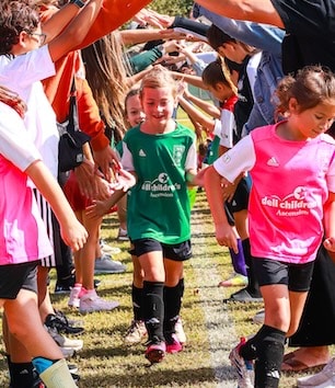 Children in sports uniforms run through a tunnel of cheering people on a sunny day at a soccer event.