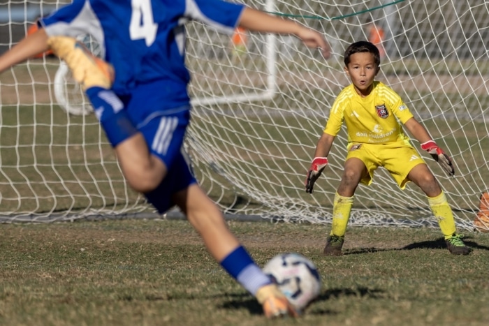 Youth soccer goalie in yellow ready to defend the goal as an opposing player kicks the ball