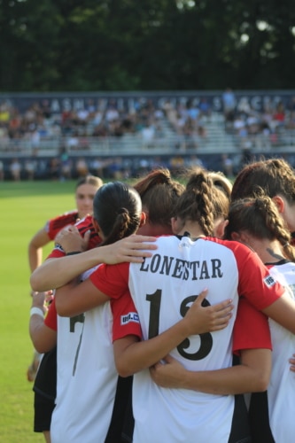 Soccer players in a team huddle on the field during a match with a blurred crowd in the background